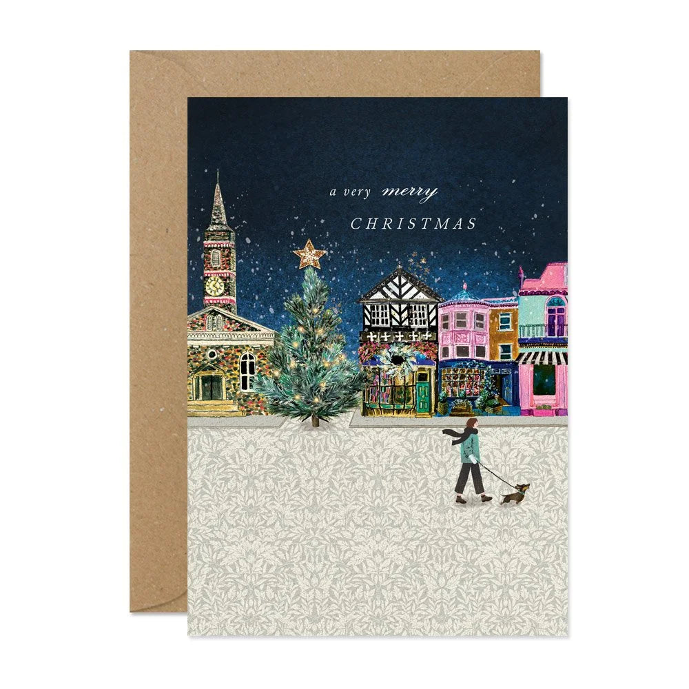 Christmas card with a snowy village scene, Christmas tree, and person with dog on a white background.