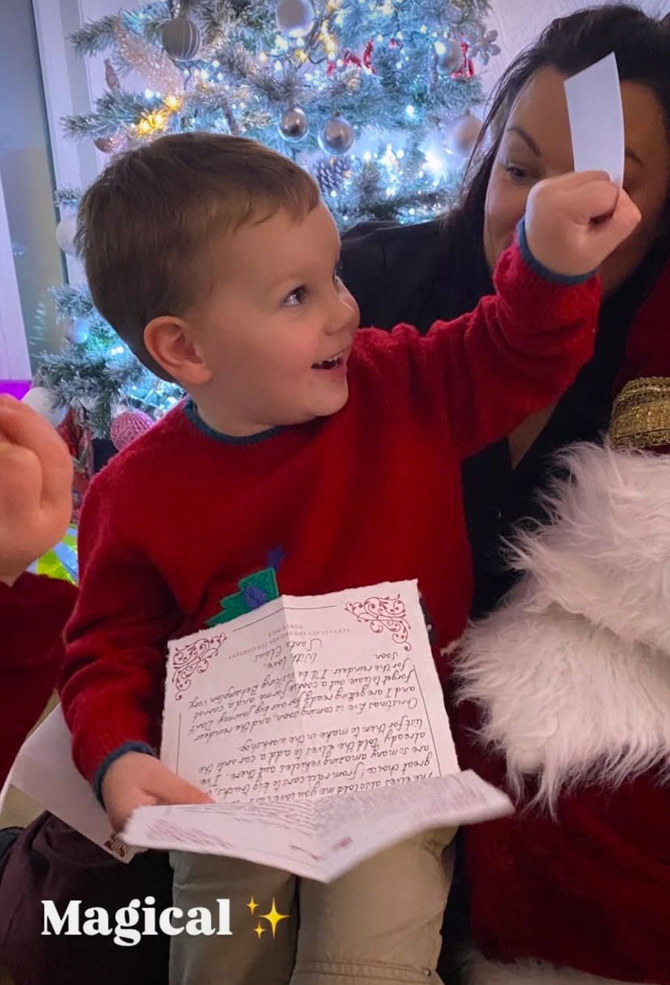 Child holding a letter with a woman and Santa Claus in front of a Christmas tree.